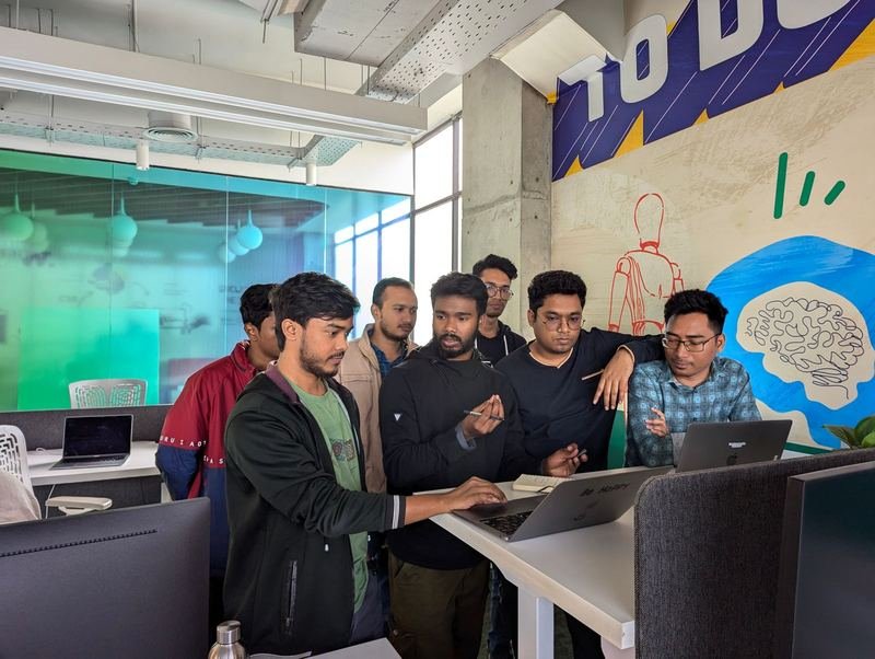 Group of young South Asian professionals gathered around a laptop at a standing desk in a modern open-plan office