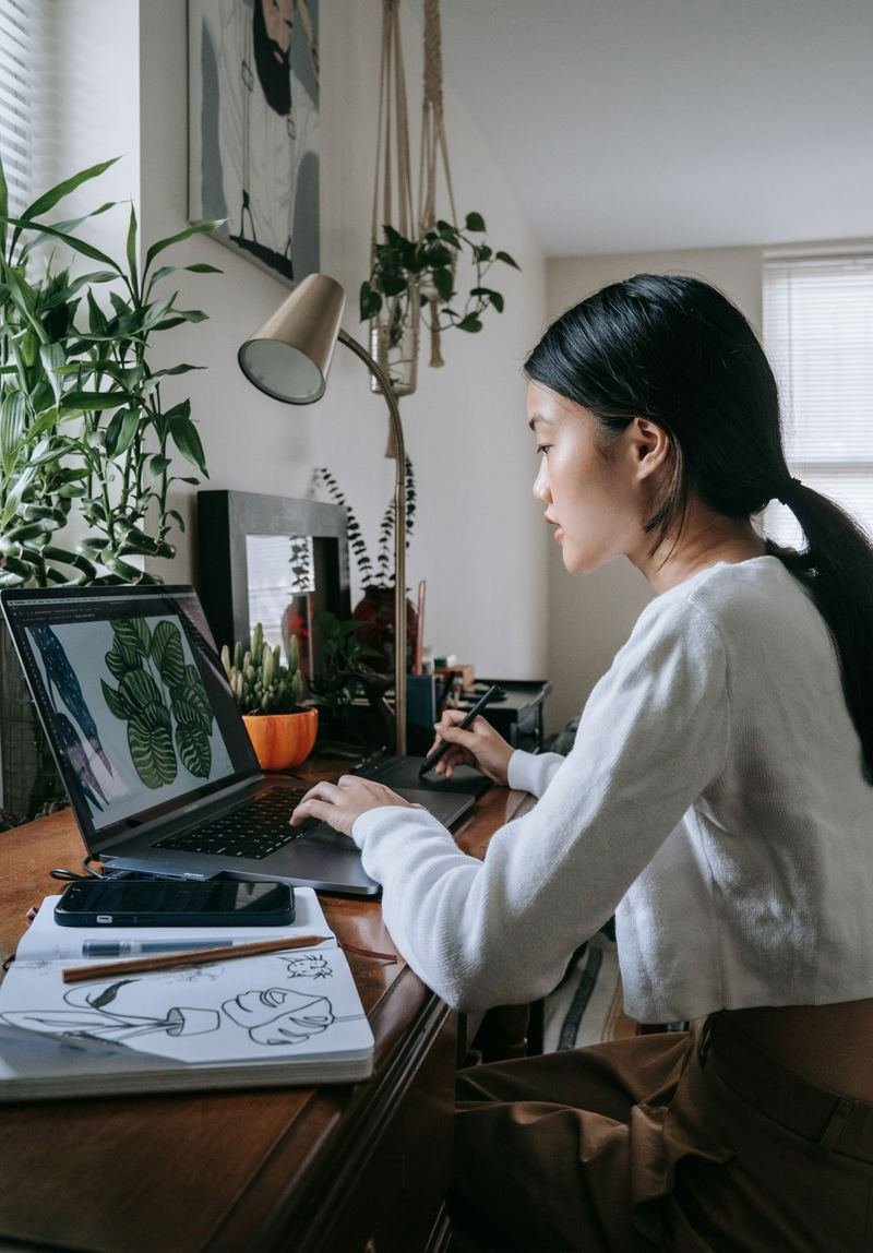 Designer working on a digital illustration on a laptop with a pen tablet, sketchbook beside her