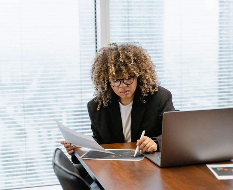Professional reviewing printed data charts next to a laptop at a desk by the window