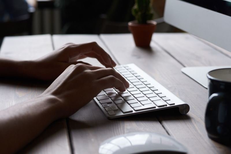 Close-up of hands typing on a white wireless keyboard on a wooden desk